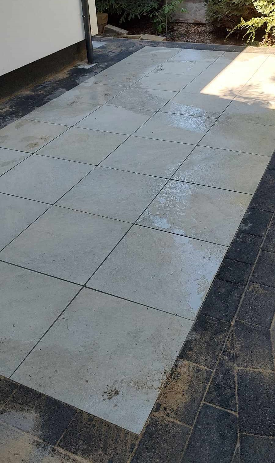 Ułożona nawierzchnia z dużych, jasnoszarych płyt z delikatną fakturą, obramowana ciemnoszarą kostką brukową. Widoczne ślady wilgoci na płytach i drobne zabrudzenia. W tle ściana budynku i zieleń.