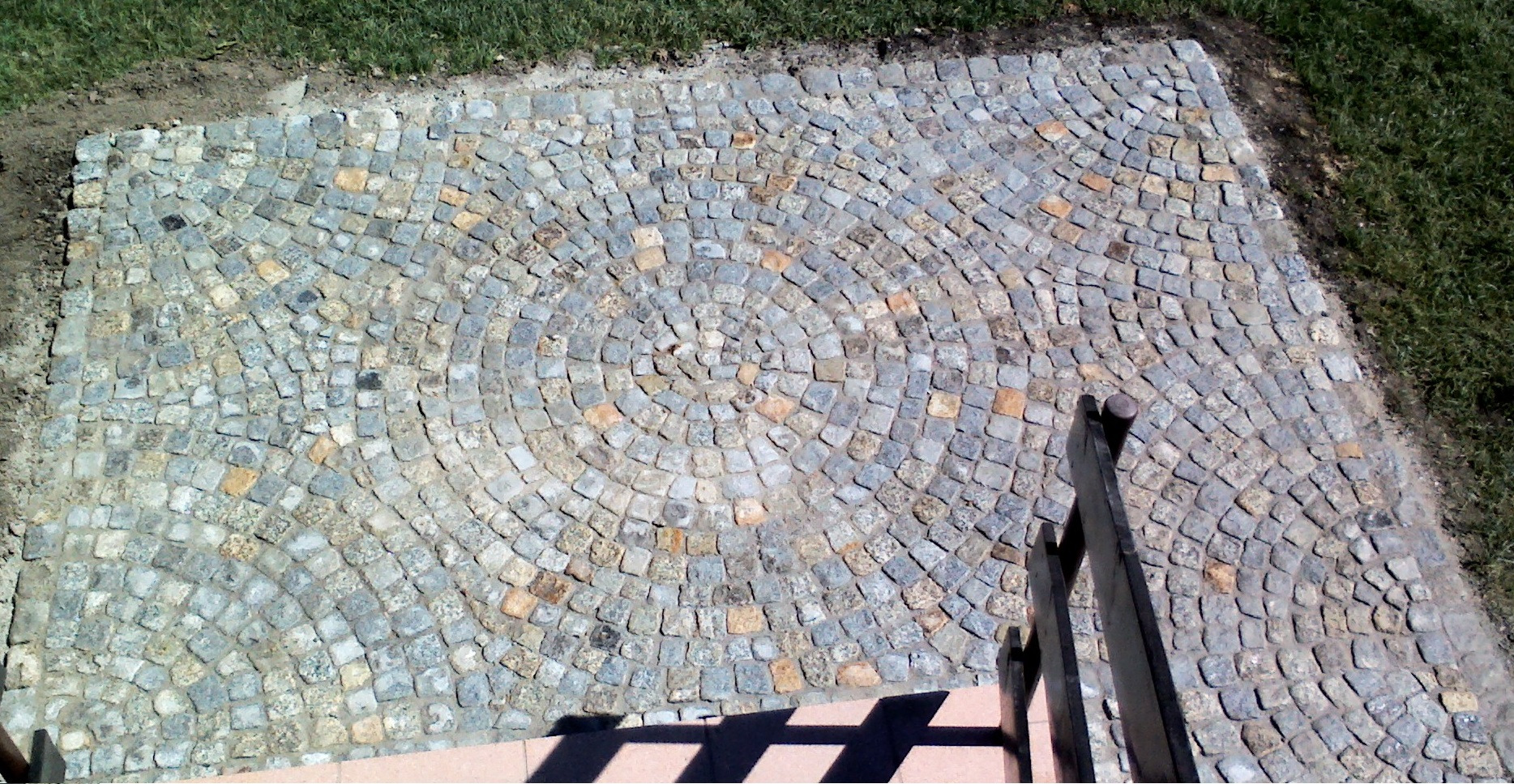 Wykończona powierzchnia z kostki granitowej ułożonej w koncentryczne okręgi, widoczne schody i trawnik w tle.