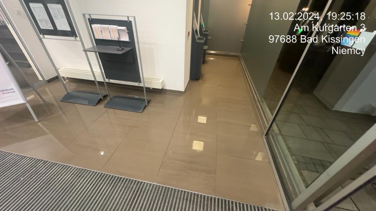 Błyszcząca, brązowa podłoga z płytek w holu wejściowym budynku, odbijająca światło, z widoczną kratką wejściową i metalowymi stojakami na ulotki.