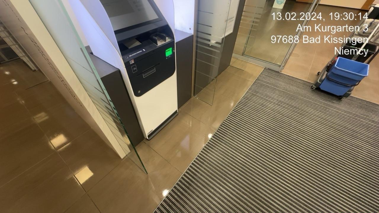 Automatyczny czytnik kart w biurze, obok wózek do mycia podłóg z niebieskim wiadrem, widoczny na podłodze z płytek i dywaniku w paski