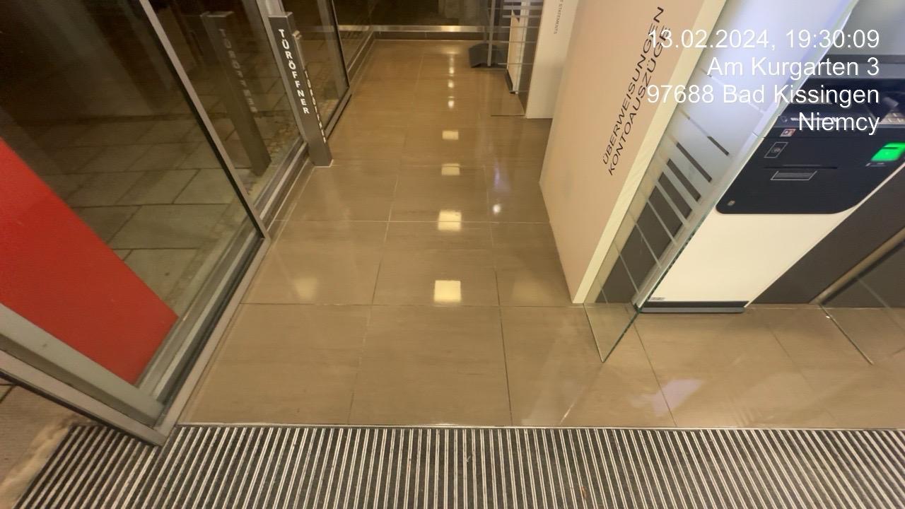 Błyszcząca, wypolerowana podłoga wewnątrz budynku, widoczna przez szklane drzwi wejściowe z metalową kratką. Na drzwiach widoczne napisy i data z godziną.