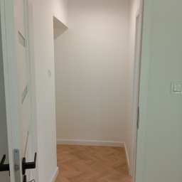 RemontyZpasją - Otwarta biała rama drzwiowa prowadząca do pustego, świeżo pomalowanego pomieszczenia z parkietem ułożonym we wzór jodełki.
