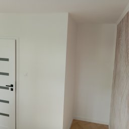 RemontyZpasją - Wnętrze pokoju po odświeżeniu: białe ściany, nowe białe drzwi z poziomymi przeszkleniami i czarną klamką, fragment ściany z tapetą w delikatny wzór liści palmowych, parkiet na podłodze.