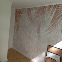 RemontyZpasją - Wnętrze pokoju z dekoracyjną ścianą z motywem palmowych liści w odcieniach beżu i brązu, parkiet na podłodze, białe listwy przypodłogowe, w rogu oparty o ścianę plakat w ramie z napisem 'La Femme'.