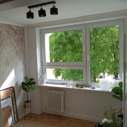 RemontyZpasją - Jasne wnętrze pokoju z wzorzystą tapetą, białym oknem z widokiem na zieleń, nowoczesną lampą sufitową i dekoracjami na parapecie.
