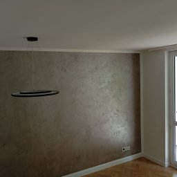 RemontyZpasją - Ściana z dekoracyjną gładzią w odcieniach brązu i szarości, podświetlona nowoczesną lampą wiszącą z dwoma okręgami, widoczne gniazdka elektryczne i parkiet w jodełkę.