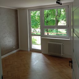 RemontyZpasją - Pusty pokój po remoncie z widokiem na zieleń za oknem balkonowym, parkiet w jodełkę, ściana pokryta dekoracyjną szarą fakturą.