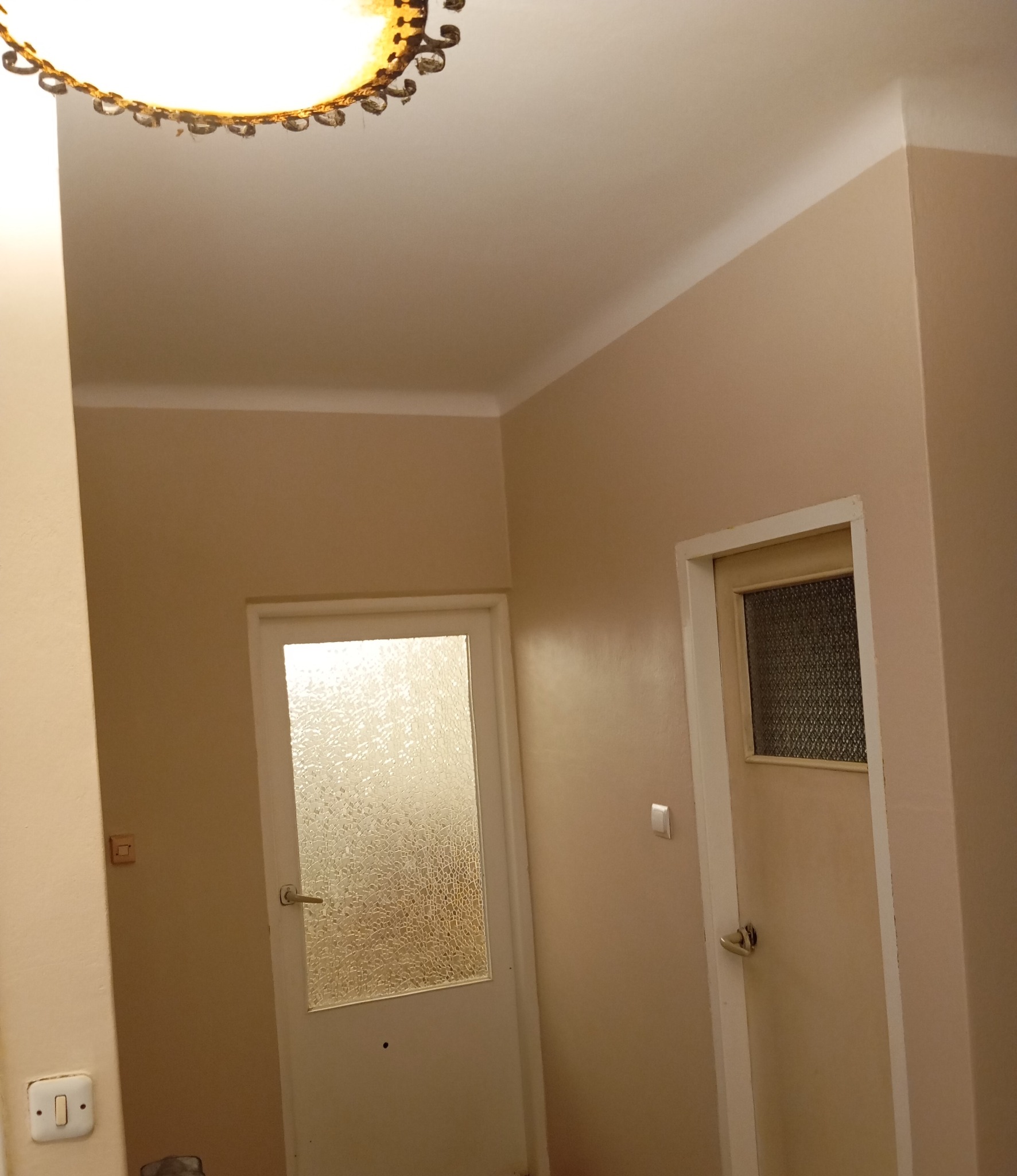 Korytarz po malowaniu na kolor beżowy, widoczne dwie białe ościeżnice drzwiowe, na suficie lampa z elementami dekoracyjnymi.