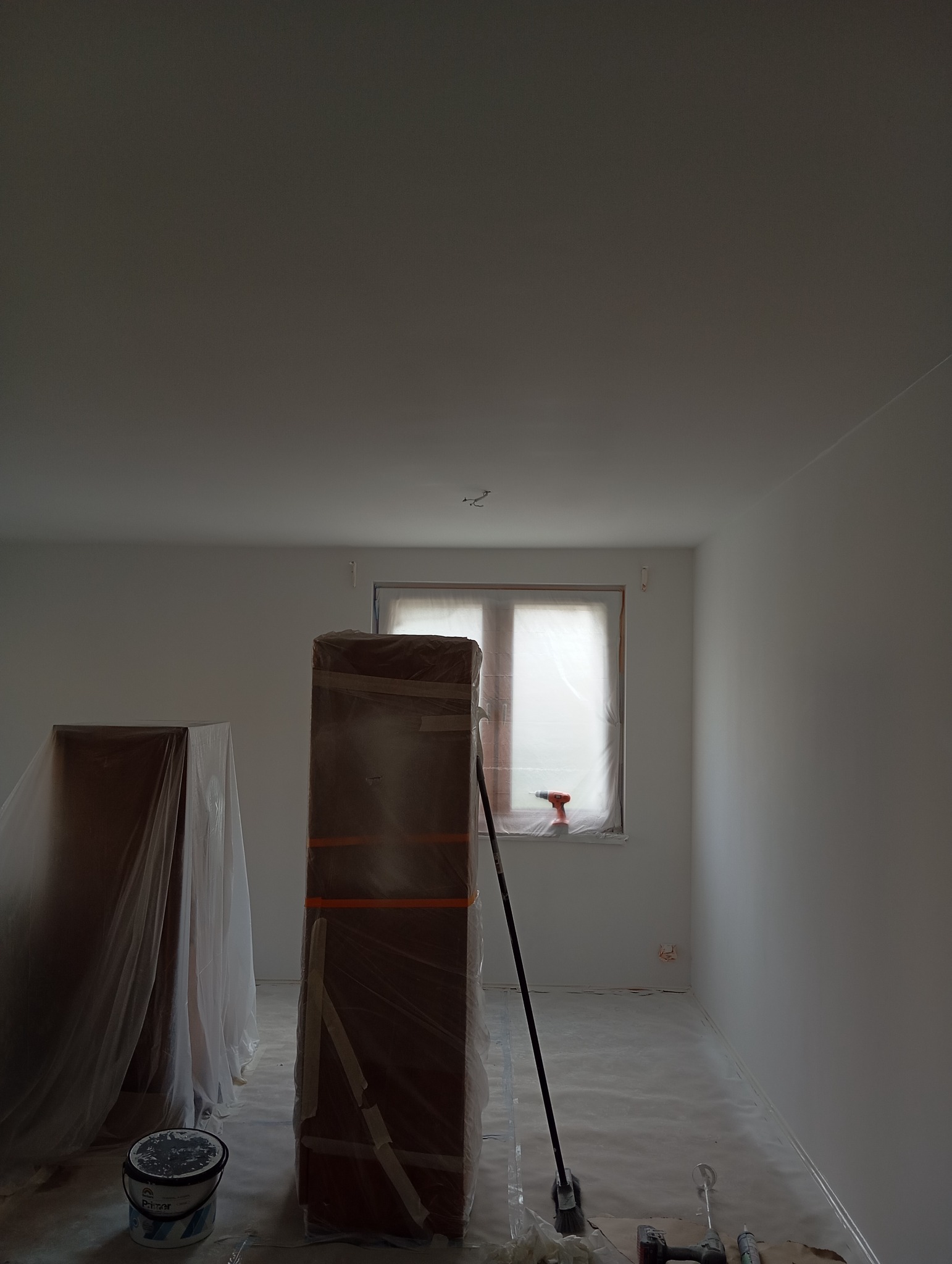 Pokój w trakcie prac wykończeniowych, zabezpieczone meble folią, świeżo pomalowane ściany, widoczne narzędzia i akcesoria malarskie.