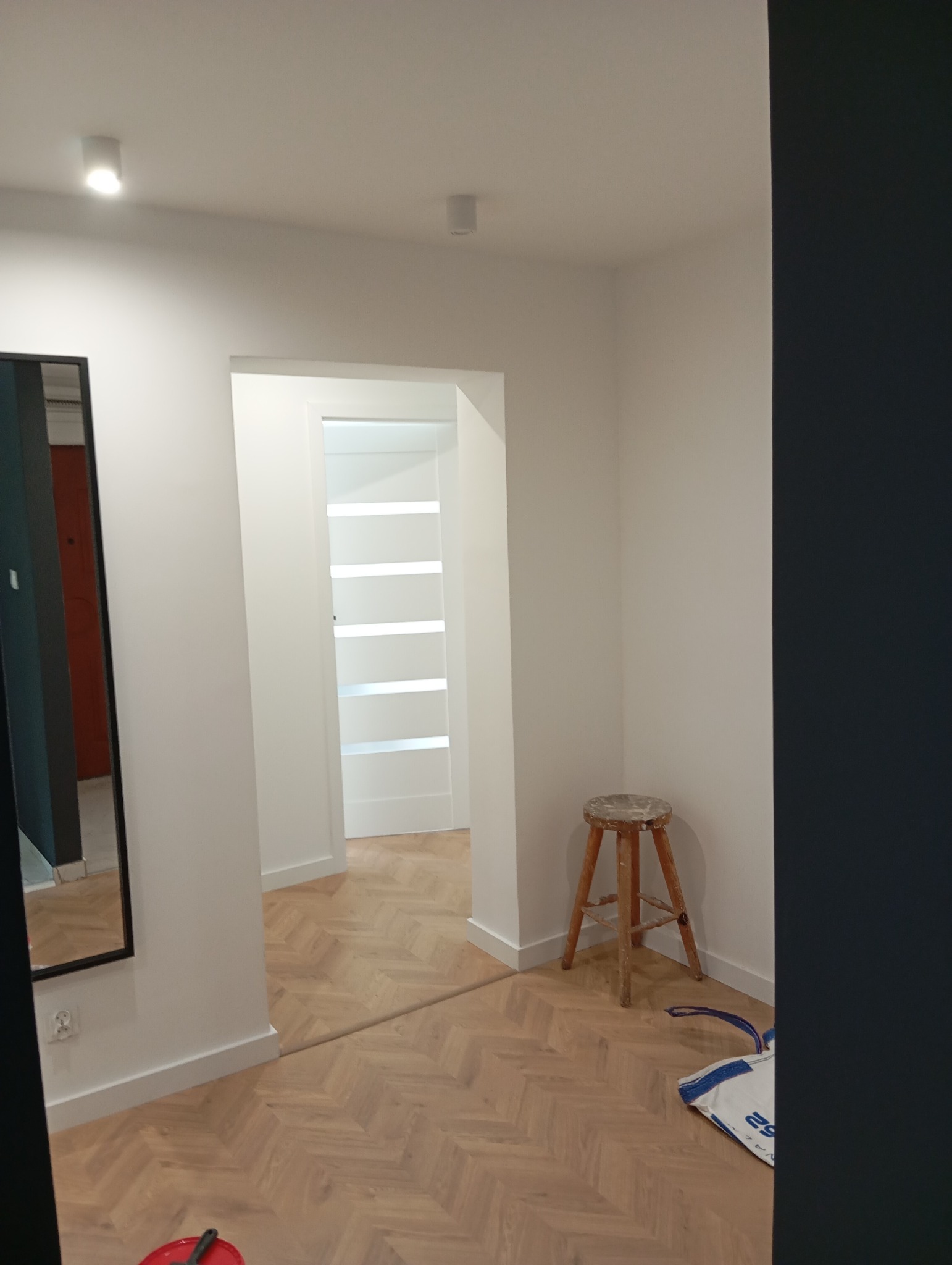 Wnętrze mieszkania po remoncie: świeżo pomalowane białe ściany, parkiet w jodełkę, białe drzwi z przeszkleniami, drewniany stołek i lustro.