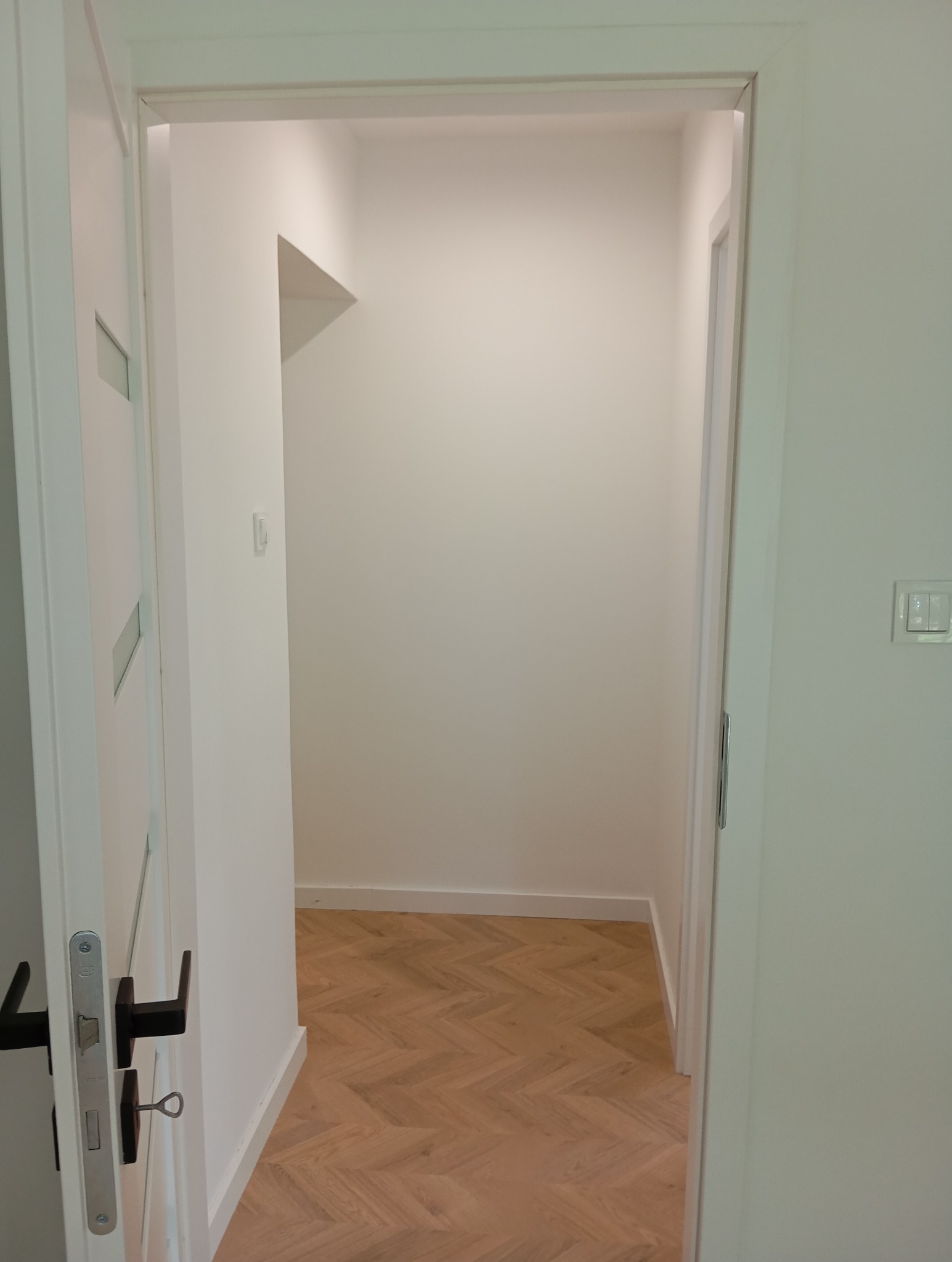 Otwarta biała rama drzwiowa prowadząca do pustego, świeżo pomalowanego pomieszczenia z parkietem ułożonym we wzór jodełki.