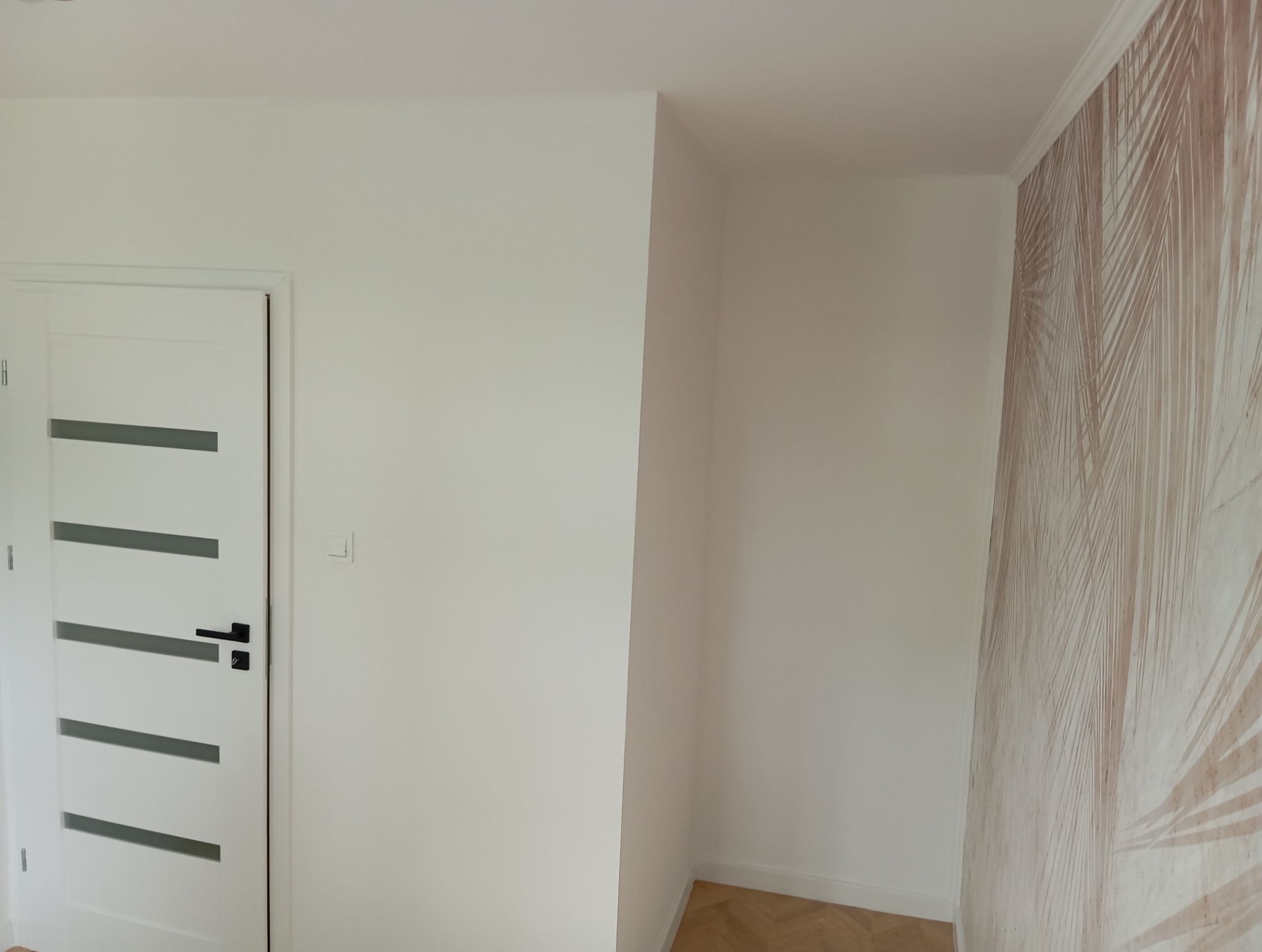 Wnętrze pokoju po odświeżeniu: białe ściany, nowe białe drzwi z poziomymi przeszkleniami i czarną klamką, fragment ściany z tapetą w delikatny wzór liści palmowych, parkiet na podłodze.