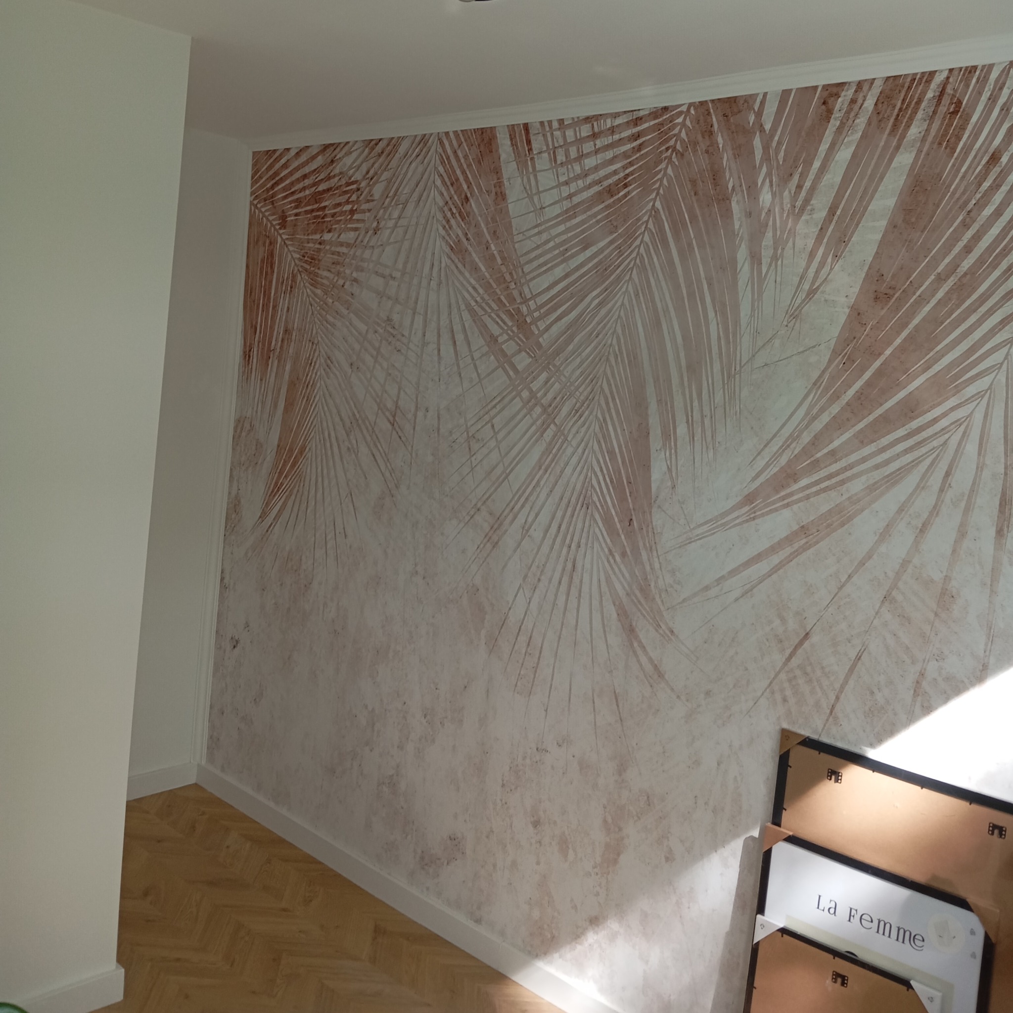 Wnętrze pokoju z dekoracyjną ścianą z motywem palmowych liści w odcieniach beżu i brązu, parkiet na podłodze, białe listwy przypodłogowe, w rogu oparty o ścianę plakat w ramie z napisem 'La Femme'.