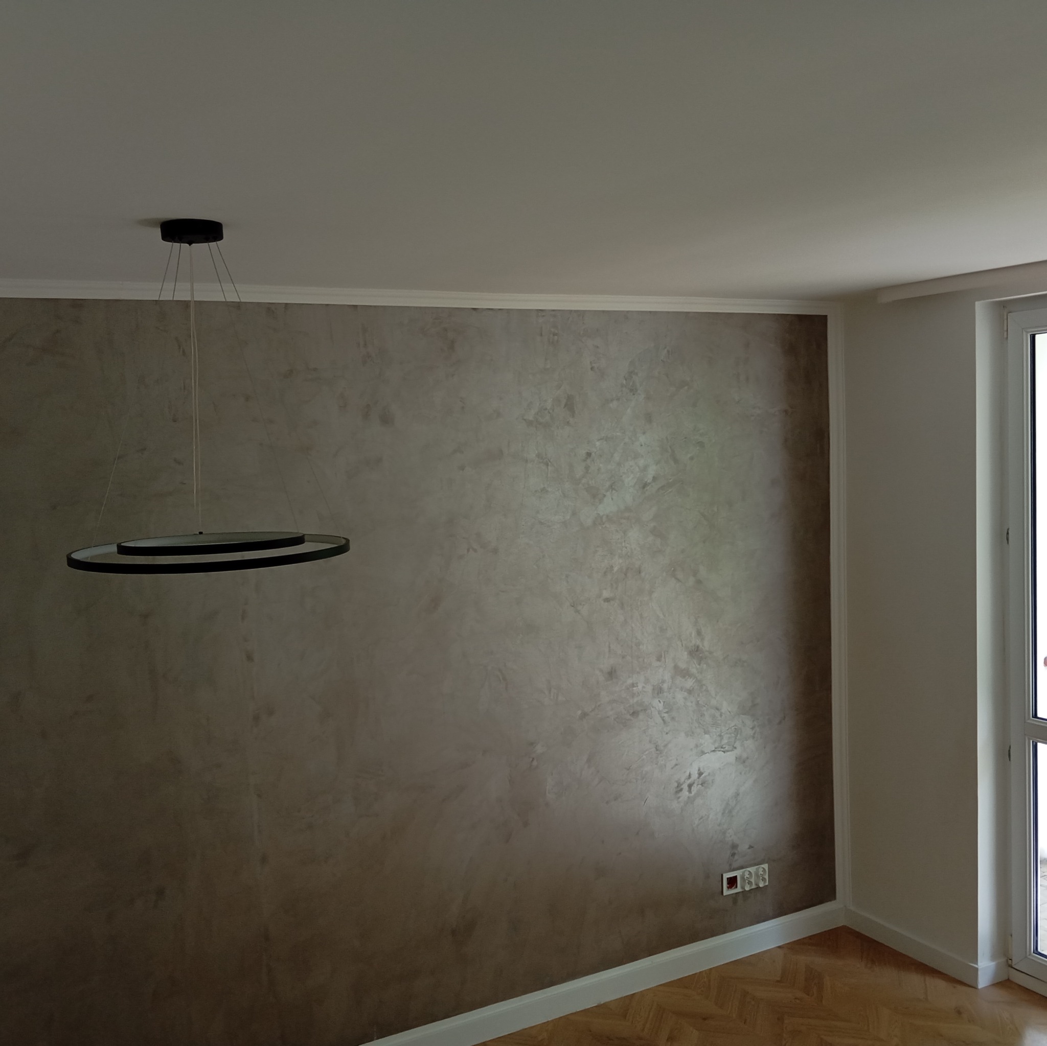 Ściana z dekoracyjną gładzią w odcieniach brązu i szarości, podświetlona nowoczesną lampą wiszącą z dwoma okręgami, widoczne gniazdka elektryczne i parkiet w jodełkę.