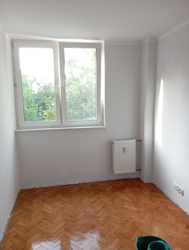 Pomalowane na biało wnętrze pokoju z parkietem ułożonym w jodełkę, widoczne ślady na podłodze i wiadro z mopem w rogu sugerujące prace wykończeniowe.