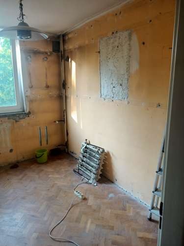 Pokój w trakcie remontu: ściany przygotowane do prac wykończeniowych, widoczne ślady po usuniętych elementach, odsłonięte fragmenty muru, podłoga z parkietu, grzejnik elektryczny podłączony...