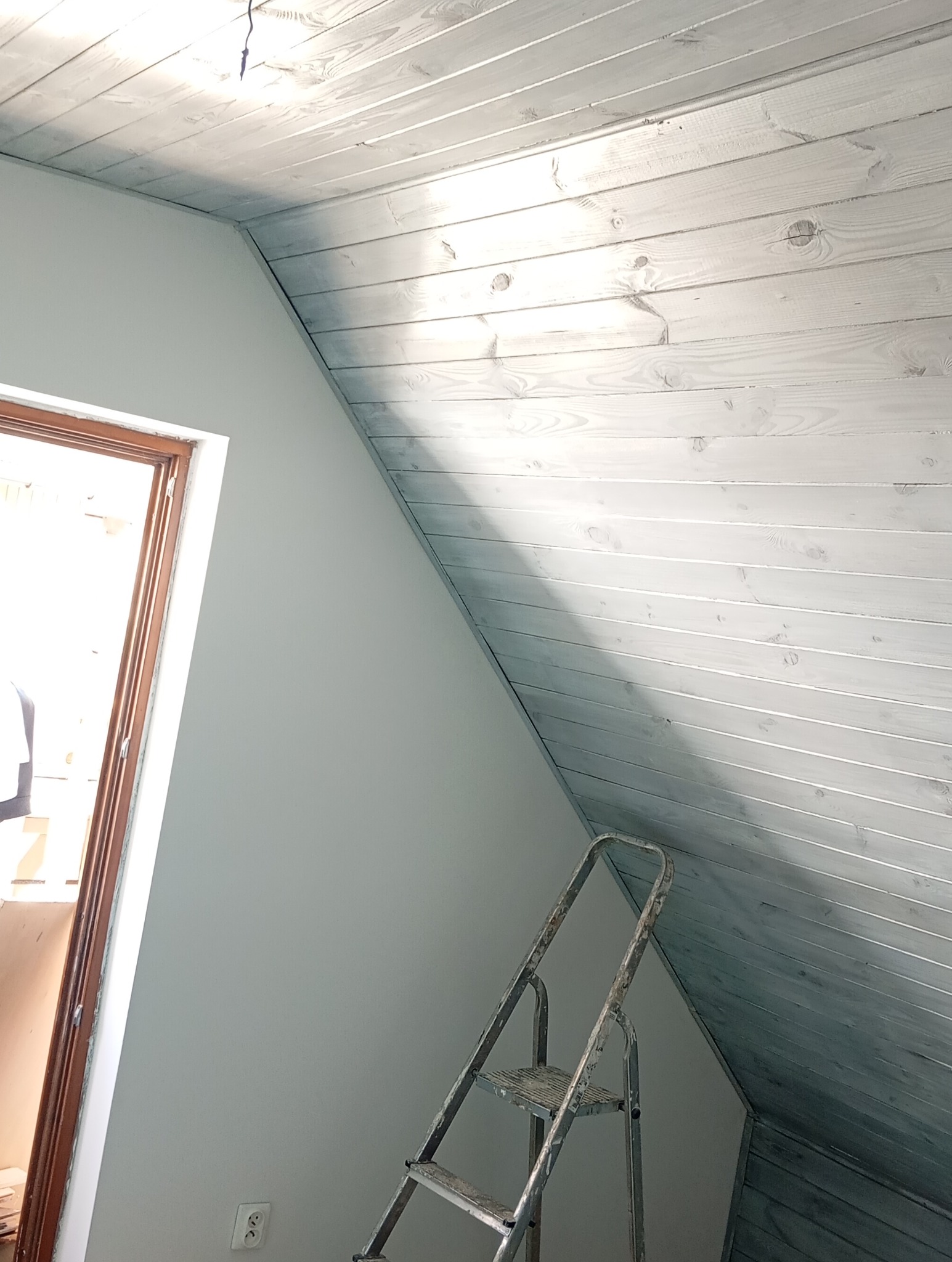 Pomalowany na szaro drewniany sufit poddasza z widoczną ścianą, ościeżnicą drzwi i częścią drabiny malarskiej.