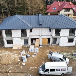 Andrzej Okrągliński ROB-DACH - Nowoczesny, dwukondygnacyjny bliźniak z szarym dachem z blachy na etapie budowy. Widoczne materiały budowlane, samochody dostawcze i osoba w kombinezonie ochronnym.