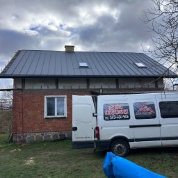 Dom z szarą blachą na dachu, częściowo ocieplony. Obok zaparkowany biały bus z reklamą usług dekarskich. Widoczne rusztowanie i elementy przygotowane do dalszych prac.