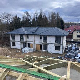 Andrzej Okrągliński ROB-DACH - Dwukondygnacyjny dom w trakcie budowy z szarym dachem, widoczna więźba dachowa i materiały budowlane. Ujęcie z góry, pochmurne niebo w tle.