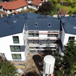 Nowoczesny budynek z ciemnym dachem z blachy na etapie wykończenia, widoczne rusztowania i elementy konstrukcyjne. Ujęcie z lotu ptaka.