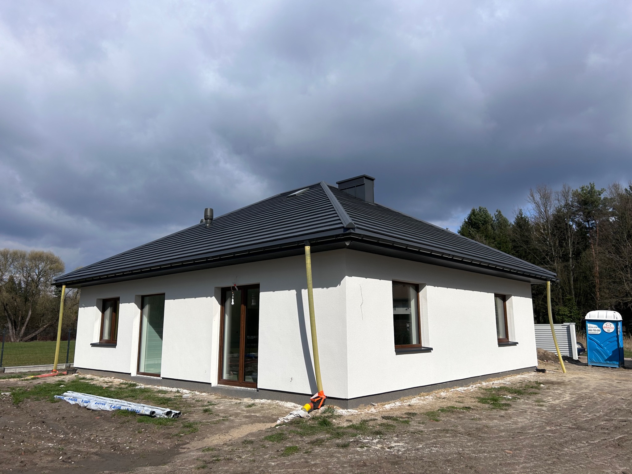 Nowoczesny dom jednorodzinny z ciemnoszarym dachem z dachówki ceramicznej, białą elewacją i widocznymi rynnami w kolorze grafitowym, na tle pochmurnego nieba i częściowo zagospodarowanego terenu.