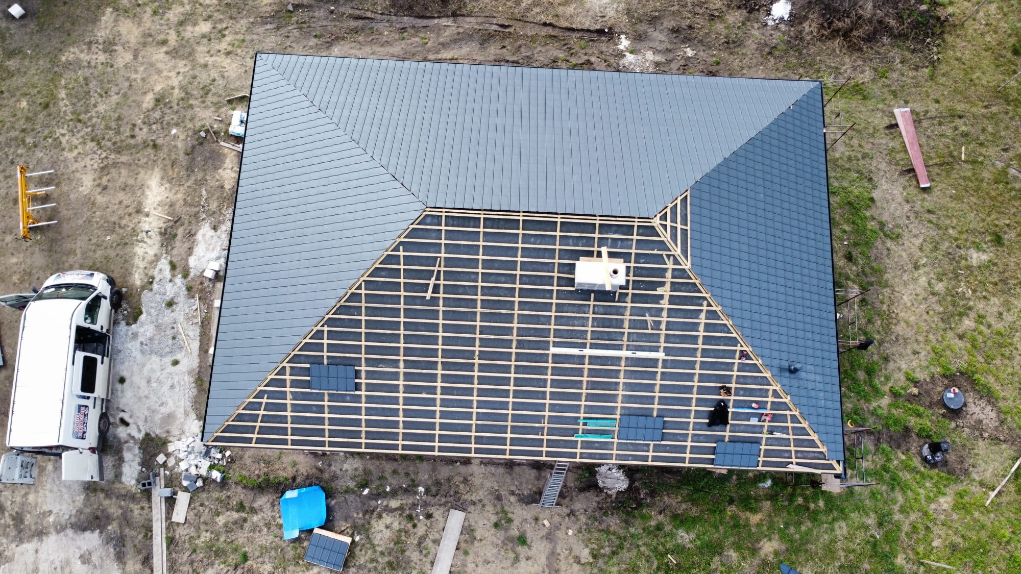 Widok z góry na dom w trakcie układania nowego pokrycia dachowego z drewnianą konstrukcją widoczną na części dachu, obok zaparkowany biały samochód dostawczy z reklamą firmy.