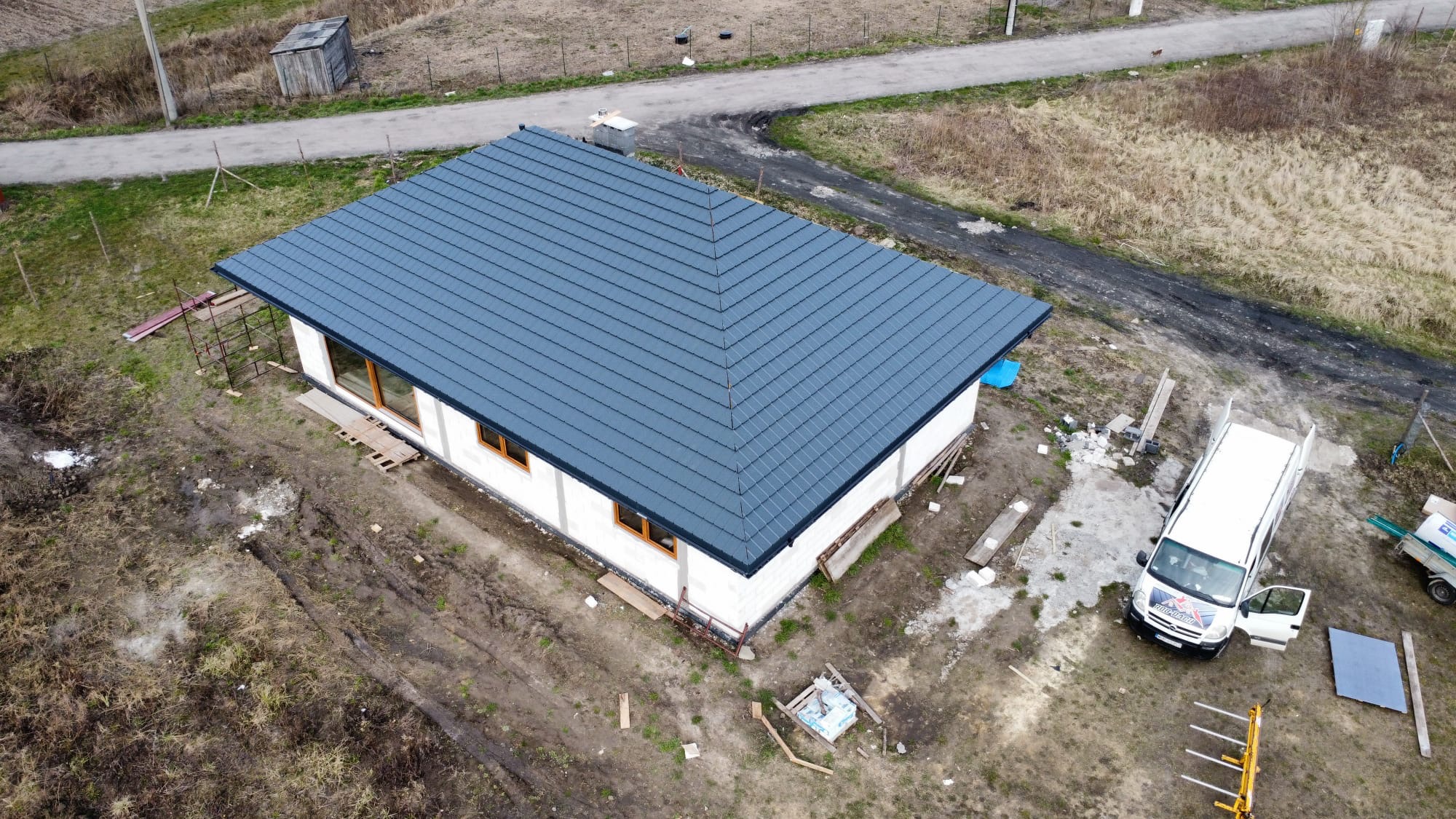 Widok z góry na dom w trakcie budowy z nowym, ciemnoszarym dachem z dachówki ceramicznej, białe ściany z pustaków, drewniane ramy okienne, rusztowanie, teren wokół domu jeszcze niezagospodarowany...