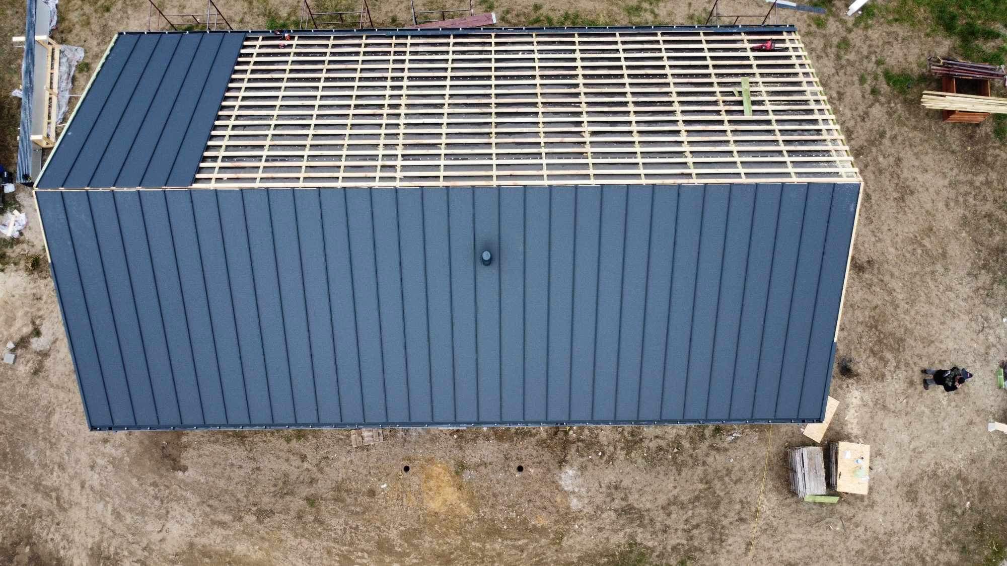 Widok z góry na częściowo pokryty, ciemnoszary blachodach na nowo budowanym domu; widoczna więźba dachowa na pozostałej części dachu.