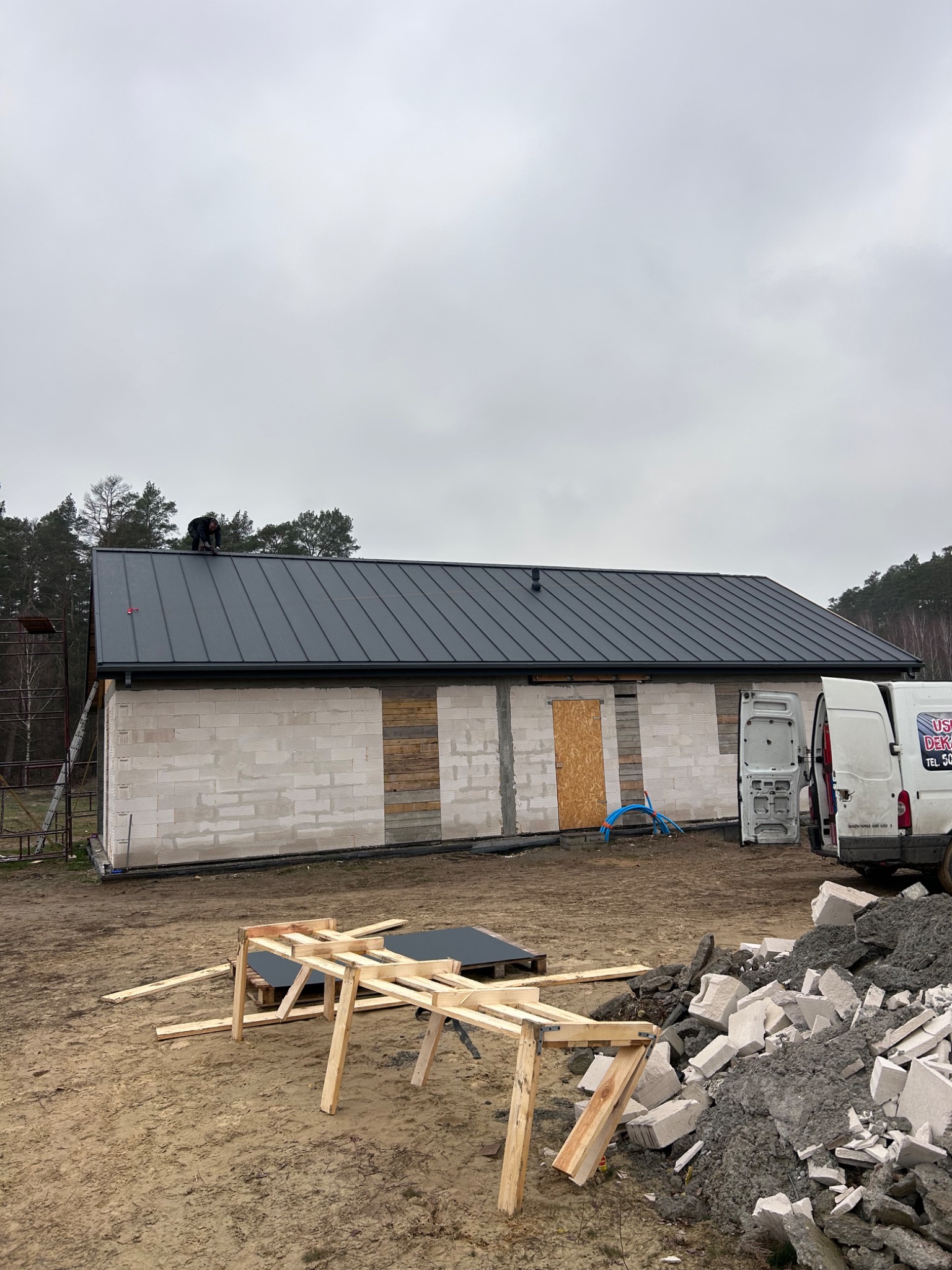 Pracownik na dachu budynku z szarej blachy trapezowej, ściany z białego bloczka, w tle las i samochód dostawczy z otwartymi drzwiami, na pierwszym planie drewniana konstrukcja i gruz.