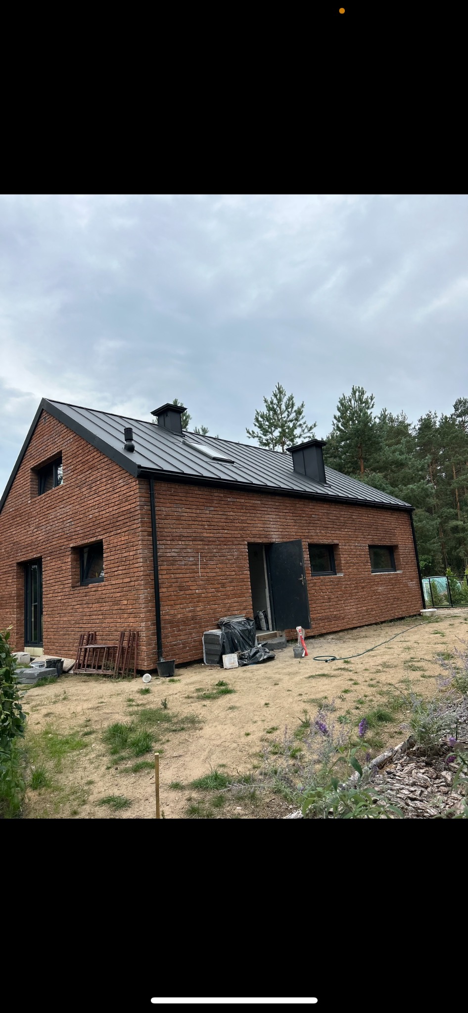 Nowoczesny dom jednorodzinny z czerwonej cegły z ciemnym dachem blaszanym i czarnymi obróbkami, widoczne materiały budowlane i rusztowanie przed budynkiem, otoczony lasem.