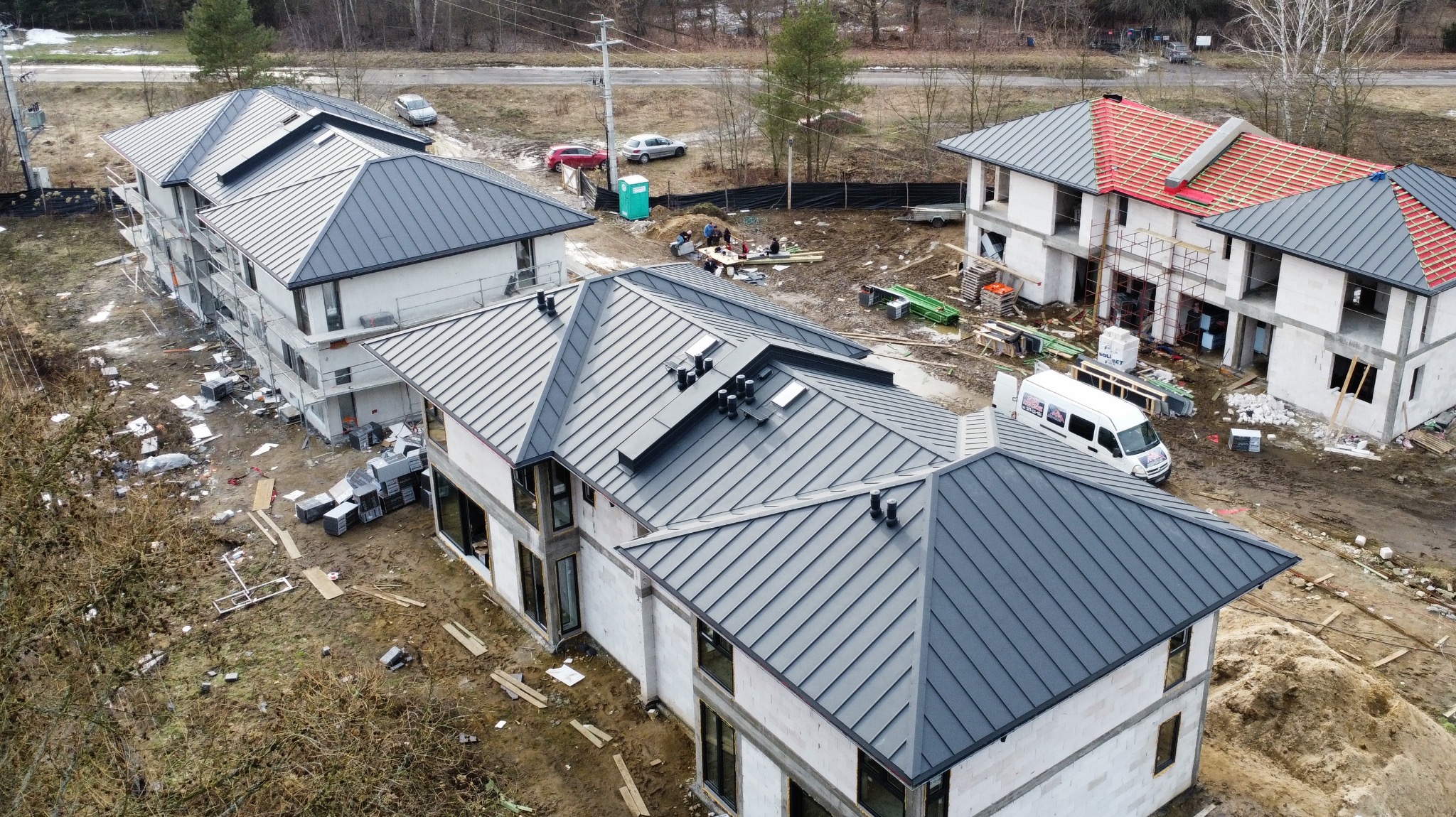 Domy w trakcie budowy z szarymi dachami z blachy na różnych etapach krycia. Widoczne rusztowania, materiały budowlane i ekipa pracująca na jednym z dachów.