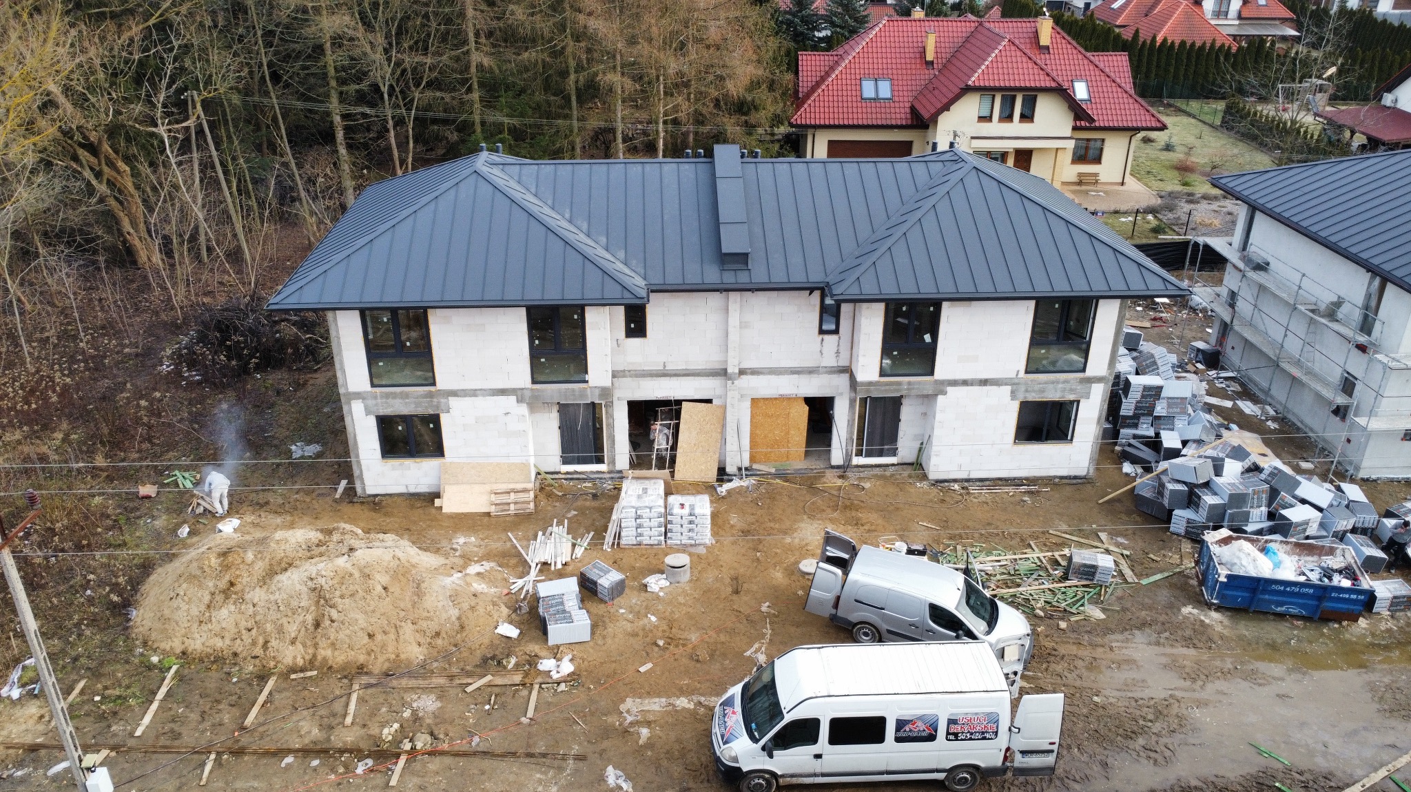 Nowoczesny, dwukondygnacyjny bliźniak z szarym dachem z blachy na etapie budowy. Widoczne materiały budowlane, samochody dostawcze i osoba w kombinezonie ochronnym.
