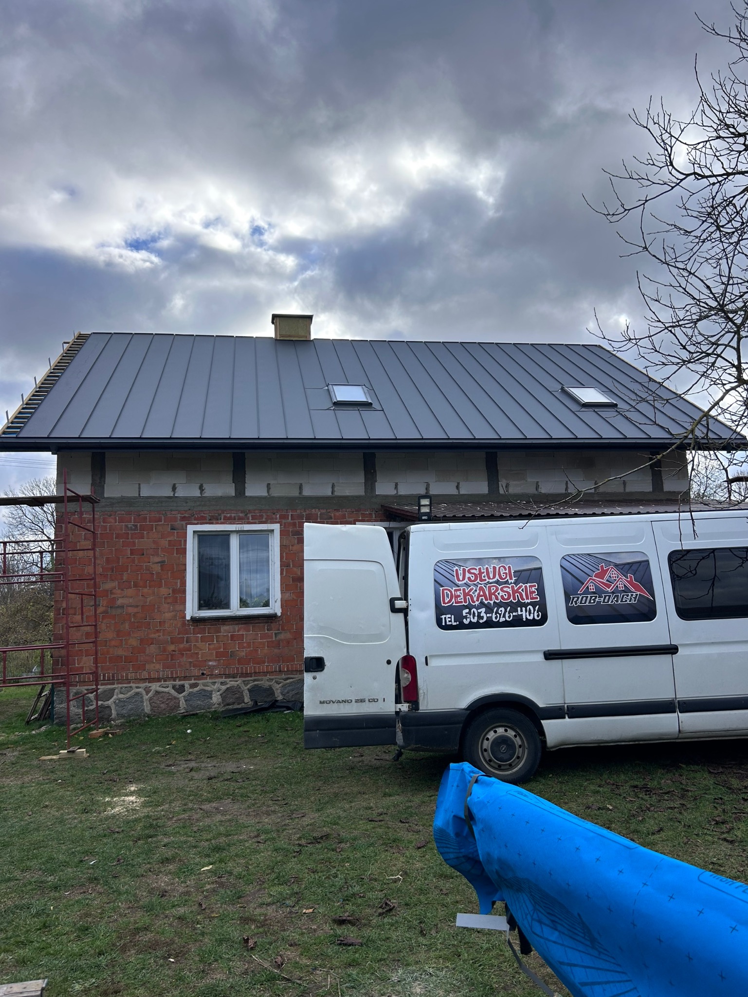 Dom z szarą blachą na dachu, częściowo ocieplony. Obok zaparkowany biały bus z reklamą usług dekarskich. Widoczne rusztowanie i elementy przygotowane do dalszych prac.