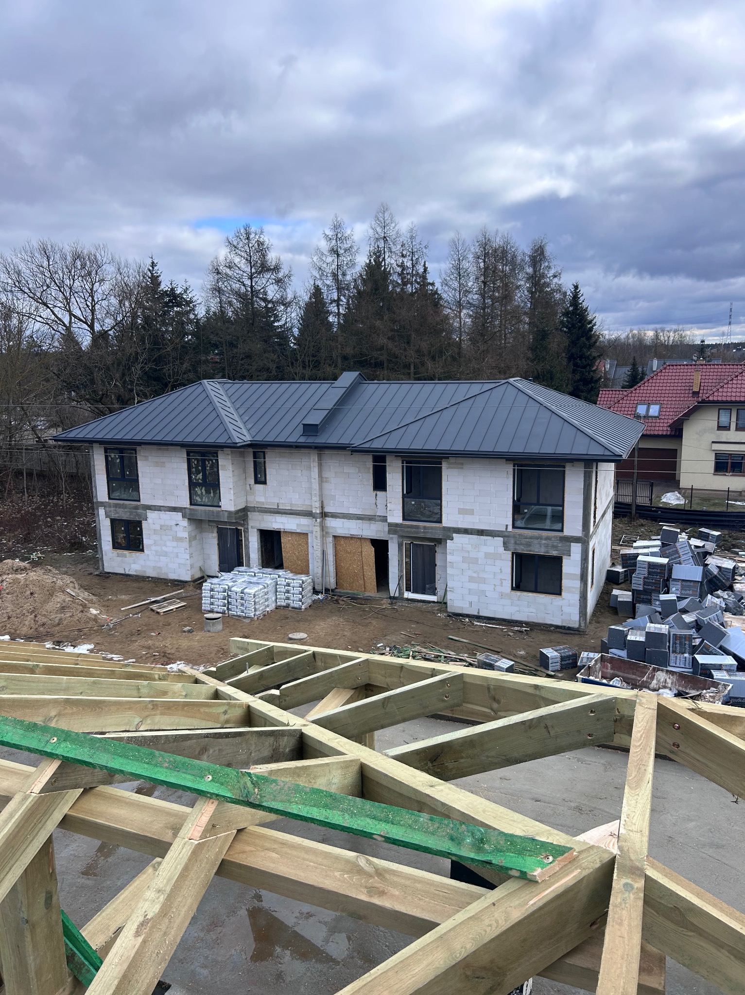 Dwukondygnacyjny dom w trakcie budowy z szarym dachem, widoczna więźba dachowa i materiały budowlane. Ujęcie z góry, pochmurne niebo w tle.