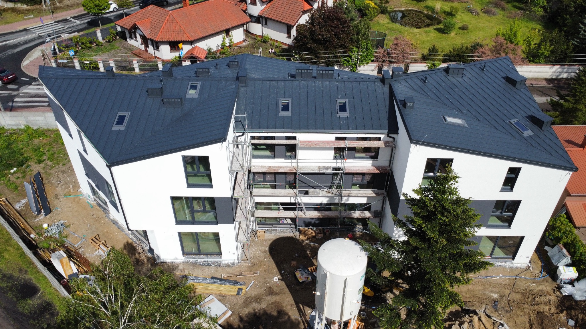 Nowoczesny budynek z ciemnym dachem z blachy na etapie wykończenia, widoczne rusztowania i elementy konstrukcyjne. Ujęcie z lotu ptaka.