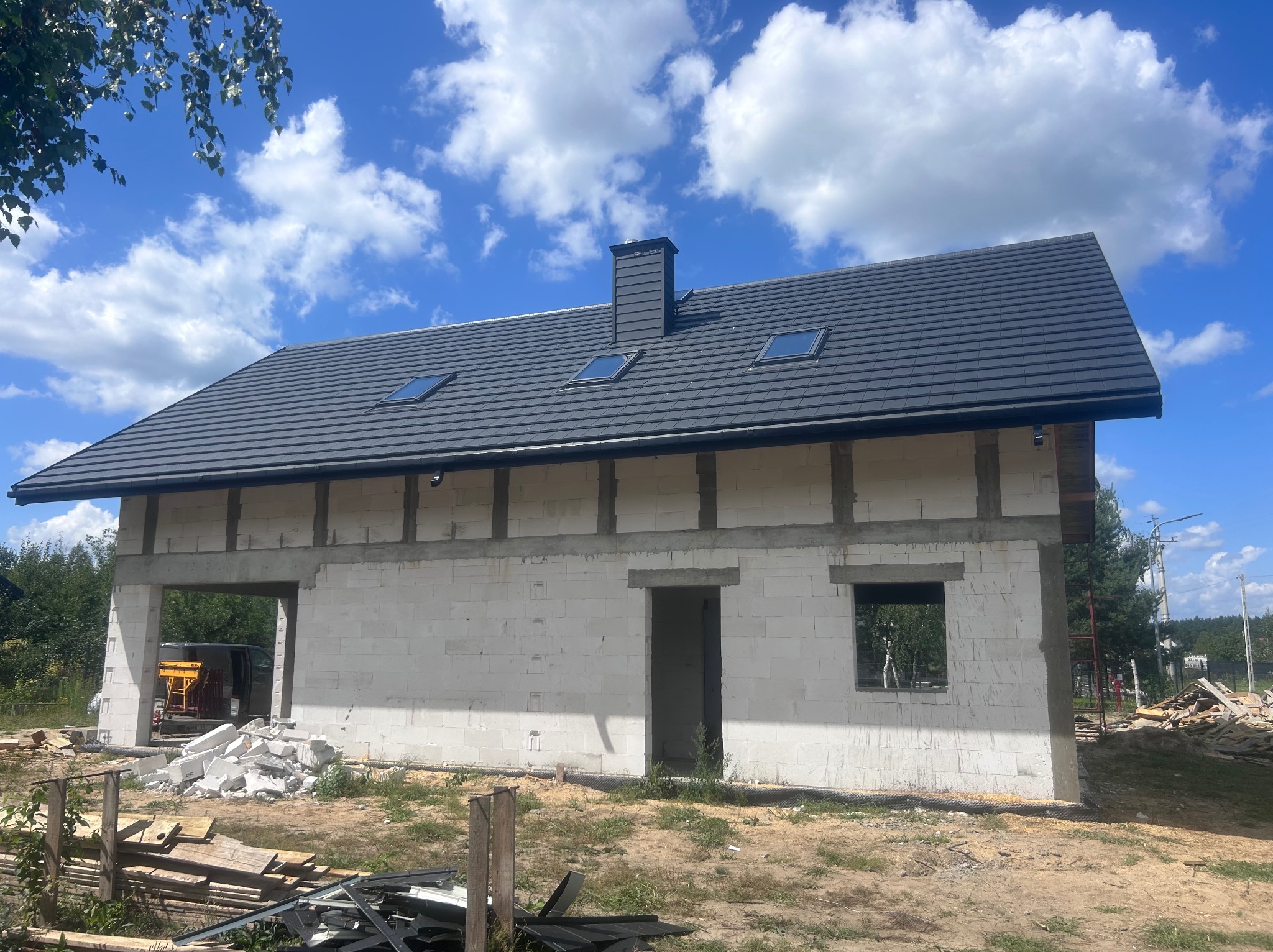 Dom w trakcie budowy z ciemnoszarą dachówką, widoczne okna dachowe i komin. Ściany z białego bloczka, otwarte przestrzenie okienne i drzwiowe. Niebo z chmurami w tle.