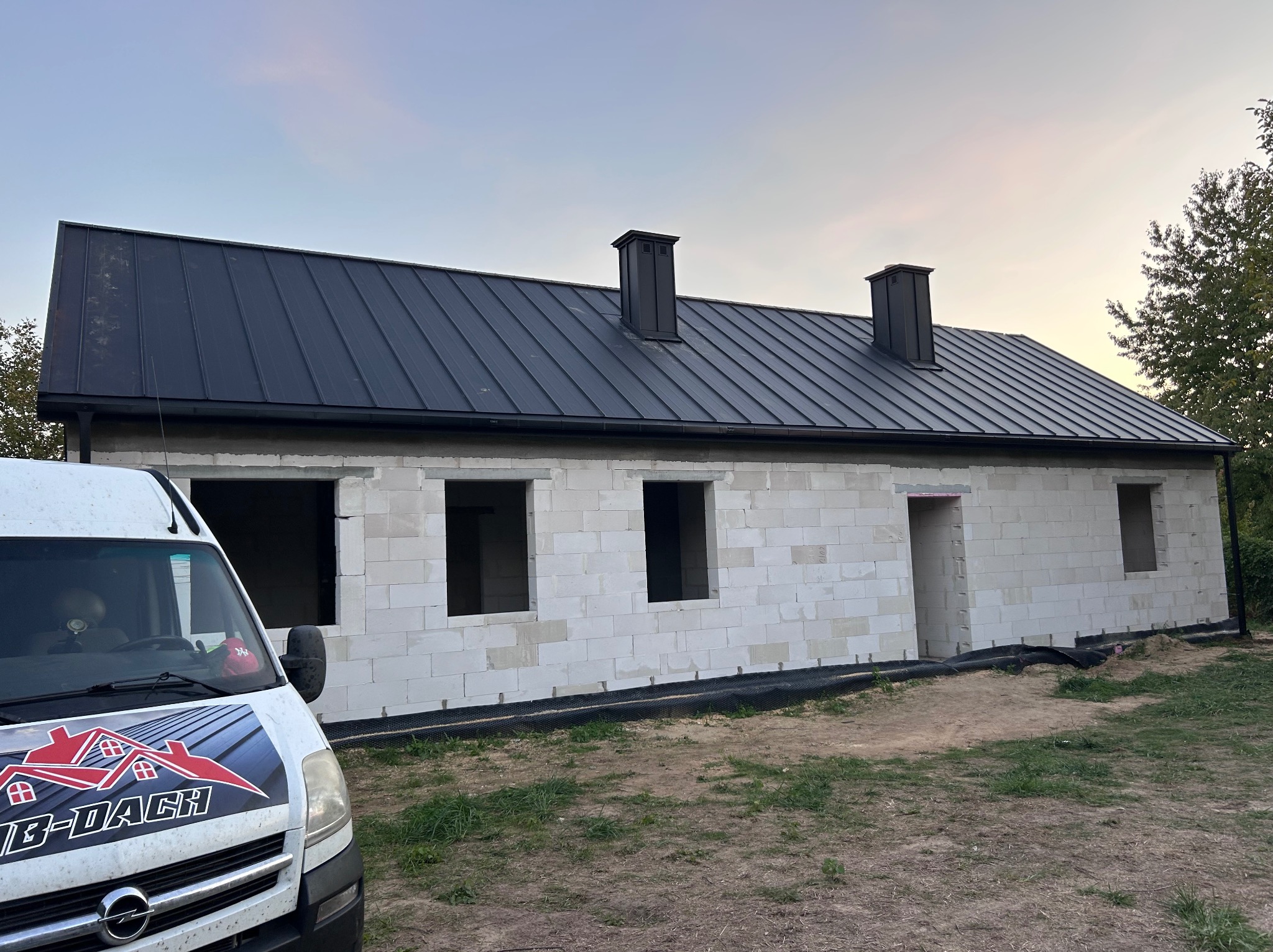Dom w trakcie budowy z czarnym dachem z blachy na rąbek stojący i dwoma kominami. Na pierwszym planie samochód dostawczy z logo firmy dekarskiej.