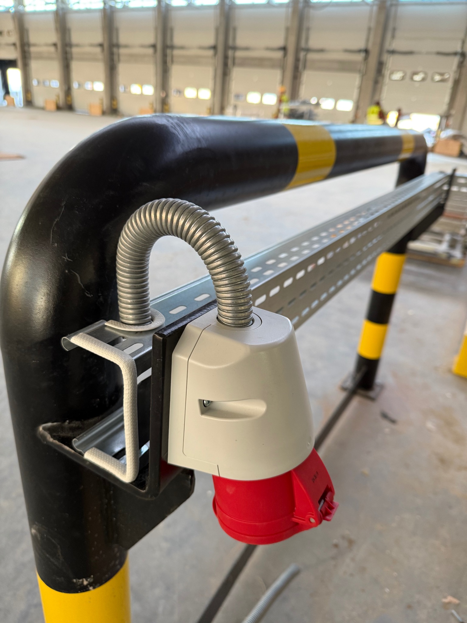 Gniazdo siłowe zamontowane na słupku ochronnym w hali magazynowej. Widoczna metalowa rurka ochronna i konstrukcja wsporcza. Słupek w kolorze czarnym z żółtymi pasami.