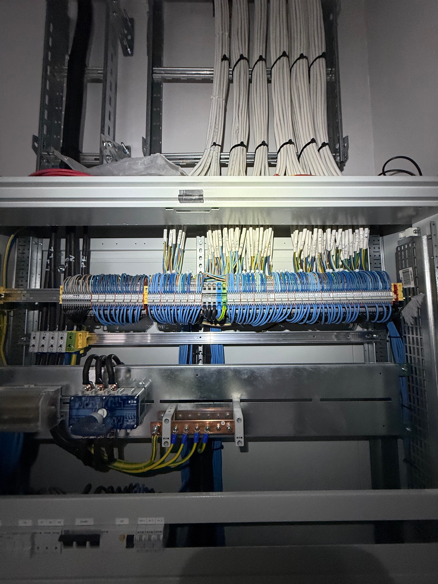 Rozdzielnia elektryczna z precyzyjnie ułożonymi kablami w kolorach białym i niebieskim, zamontowanymi na szynach DIN. Widoczne elementy sterownicze i zabezpieczenia.