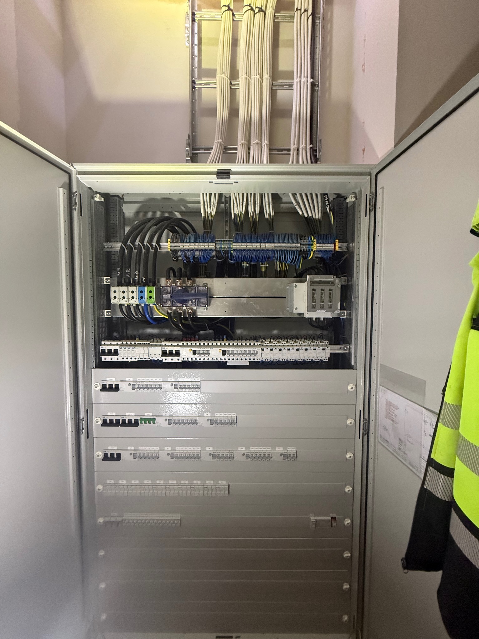 Otwarta szafa elektryczna z precyzyjnie ułożonymi kablami i elementami sterującymi. Widoczne przewody w korytkach nad szafą oraz schemat na drzwiach.