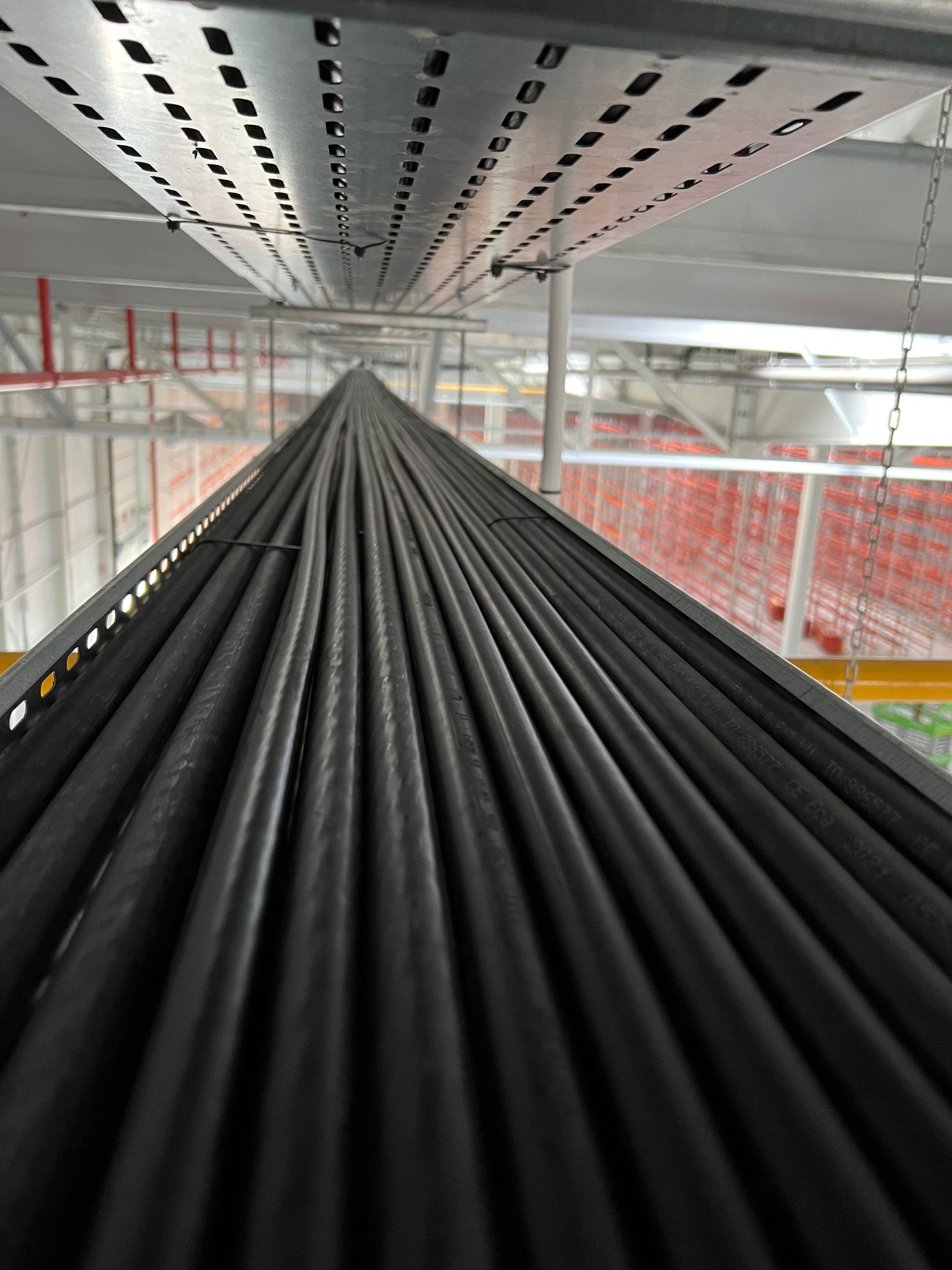 Rząd ułożonych kabli elektrycznych na metalowej konstrukcji wewnątrz budynku, widok z dołu, perspektywa zbieżna, oświetlenie górne.