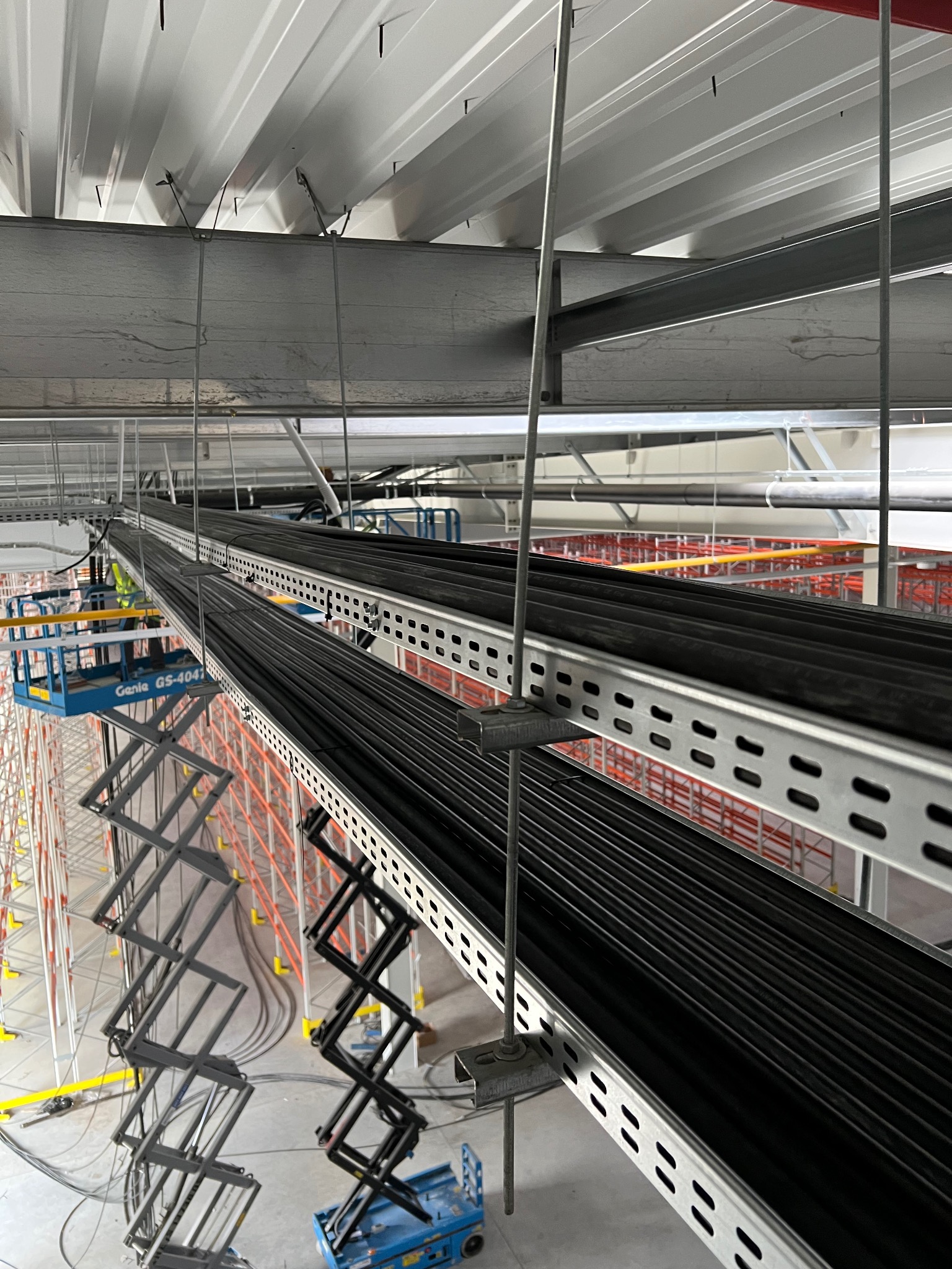 Montaż tras kablowych pod sufitem hali magazynowej, widok z podnośnika koszowego. Metalowe korytka z kablami, szare ściany i sufit. Prace instalacyjne w toku.
