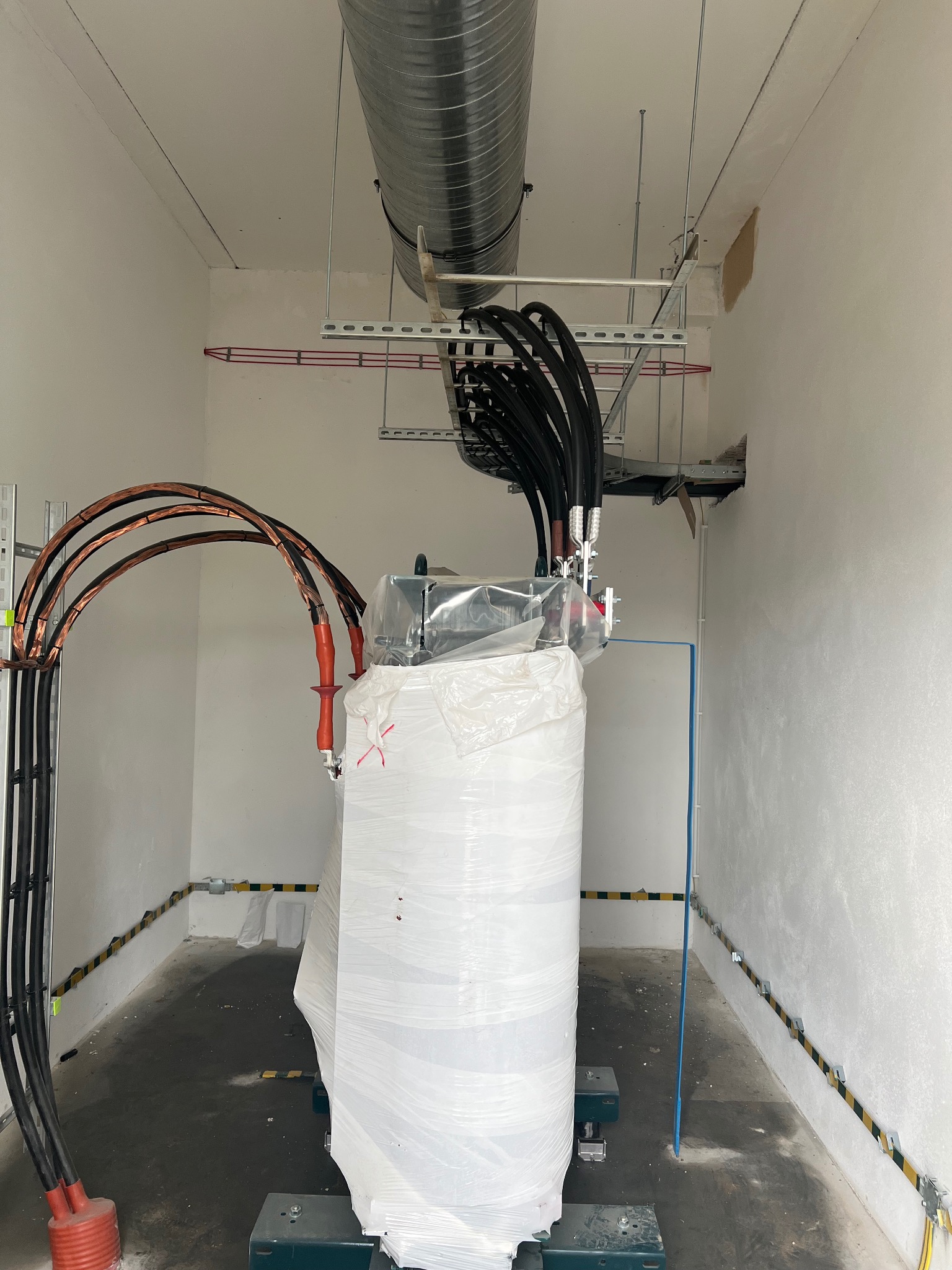 Transformator w folii ochronnej w pomieszczeniu z instalacją elektryczną, widoczne kable, rury wentylacyjne i elementy montażowe na ścianach.