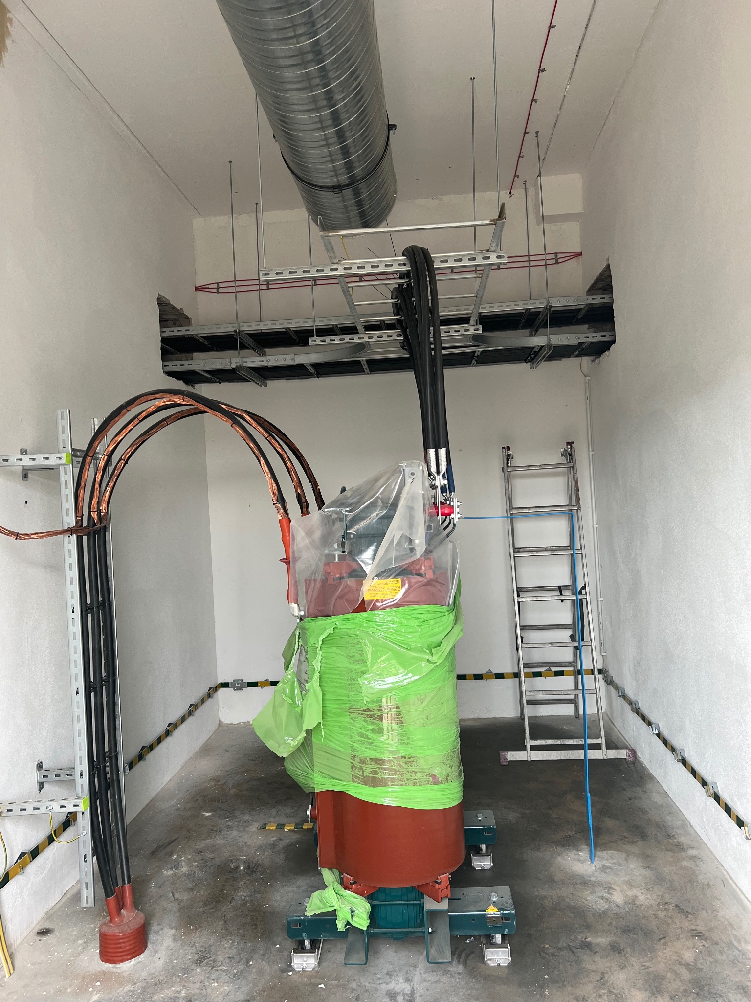 Transformator w pomieszczeniu technicznym, zabezpieczony folią, widoczne kable elektryczne, drabina i metalowe korytka kablowe pod sufitem.