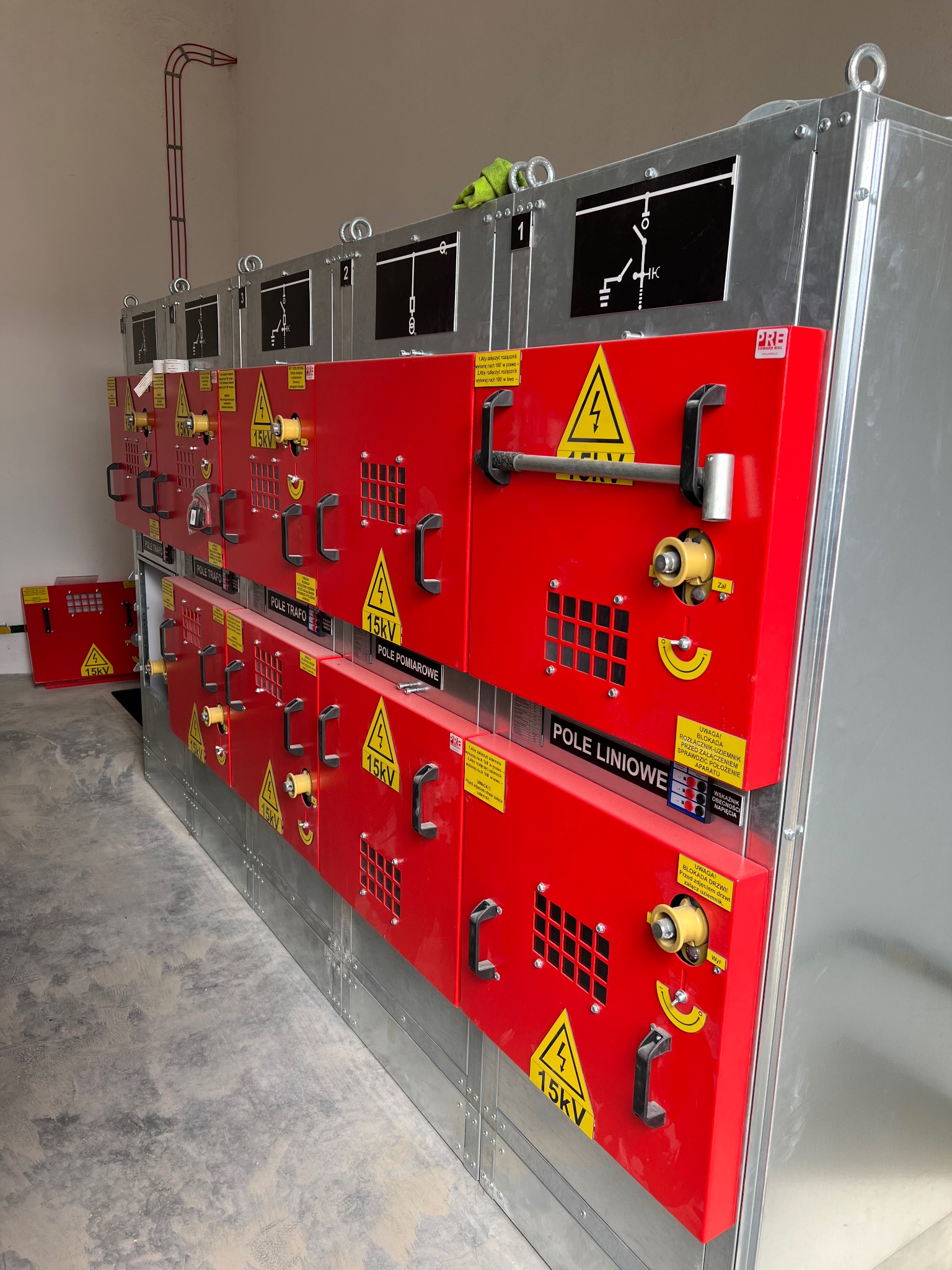 Rozdzielnica elektryczna 15kV z czerwonymi panelami i oznaczeniami ostrzegawczymi. Widoczne schematy i elementy sterujące. Instalacja w pomieszczeniu technicznym.