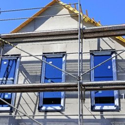 JP TANI DOM SPÓŁKA Z OGRANICZONĄ ODPOWIEDZIALNOŚCIĄ - Dom w trakcie ocieplania, widoczne rusztowania, okna z niebieskimi refleksami i fragment dachu z żółtą folią izolacyjną, data i godzina na zdjęciu.