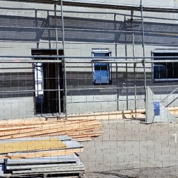 JP TANI DOM SPÓŁKA Z OGRANICZONĄ ODPOWIEDZIALNOŚCIĄ - Surowy stan domu w trakcie budowy, widoczne rusztowania, przygotowane deski, elewacja w trakcie ocieplania z widocznymi otworami okiennymi i wejściowymi.