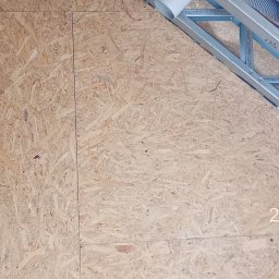 JP TANI DOM SPÓŁKA Z OGRANICZONĄ ODPOWIEDZIALNOŚCIĄ - Wnętrze w trakcie budowy: ściana z płyty OSB na metalowym stelażu, widoczne elementy konstrukcji dachu i rury wentylacyjne, żółta miarka na dole po lewej stronie.