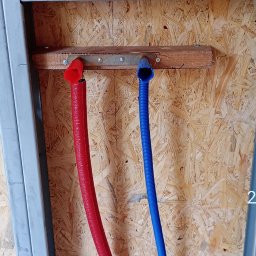 JP TANI DOM SPÓŁKA Z OGRANICZONĄ ODPOWIEDZIALNOŚCIĄ - Czerwona i niebieska rura wodna przymocowane do drewnianej belki na ścianie z płyty OSB, widoczne metalowe profile konstrukcyjne.